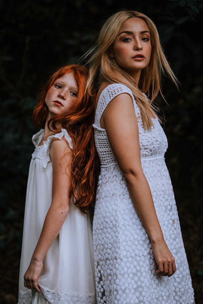 A mother and daughter posing gracefully in elegant white dresses outdoors.